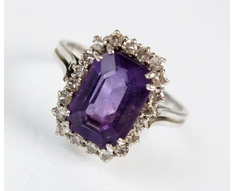 An amethyst and diamond cluster ring, designed as a central trap cut amethyst claw set in white metal within a border of eigh