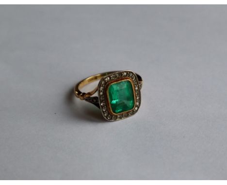 An emerald and diamond ring, the step cut emerald approximately 9mm x 8mm, surrounded by diamond chips to a white metal setti