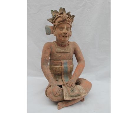 A pre-Columbian type figure seated cross legged, the hat adorned with an animal mask and arms, beaded necklace, with sash in 
