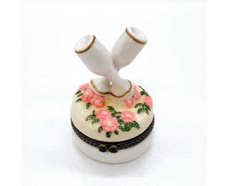 In white with pink floral and green leaf design and two champagne glasses as finial. Hinged lid with metal elements in bronze