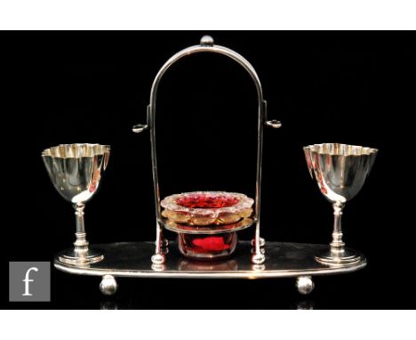 An oval plated egg cup stand raised on ball feet with two egg cups and a central carrying handle with a ruby glass open salt 
