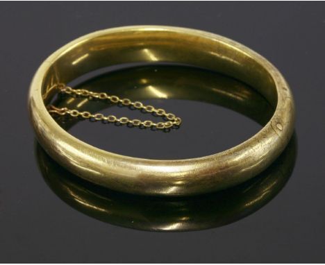 An early 20th century oval section, hinged, hollow gold bangle,with concealed box clasp and safety chain, tested as approxima