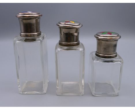 Three silver topped glass scent bottles, each lid set with enamel decoration. 10.5 cm high, 9 cm high and 8 cm high.