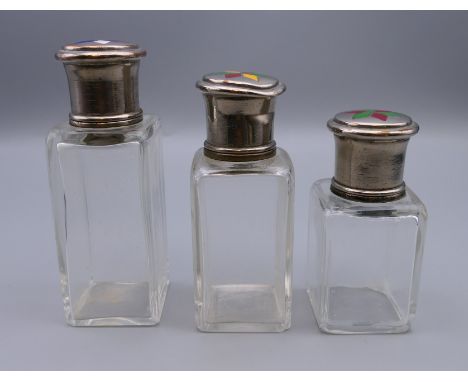 Three silver topped glass scent bottles, each lid set with enamel decoration. 10.5 cm high, 9 cm high and 8 cm high.