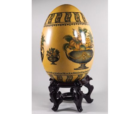 A large ceramic decorative egg with scenes of fruit in urns, raised on a five legged hard wood stand, 35cm high  