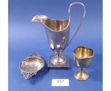 A helmet form white metal cream jug unmarked, a silver egg cup, 85g and a silver tea strainer, 35g 