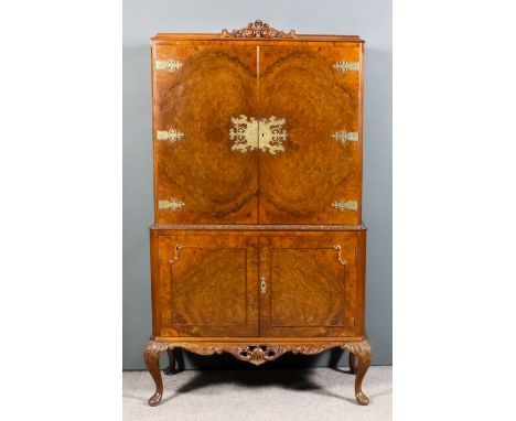 An early 20th Century figured walnut two-tier cocktail cabinet of "Queen Anne" design, the upper part with scroll carved cres