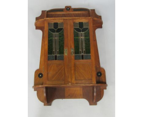 An Arts and Crafts mahogany and ebony inlaid wall mounted cupboard, the shelf top over a pair of stained glass doors, enclosi