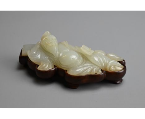 A CHINESE WHITE JADE FIGURE OF A SCHOLAR ON WOODEN STAND, QING DYNASTY. The bearded scholar resting on a pillow holding a scr