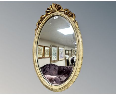 An oval-shaped bevel-edged mirror, in cream and gilt frame with ornate ribbon finial above, overall framed dimensions 75 cm x
