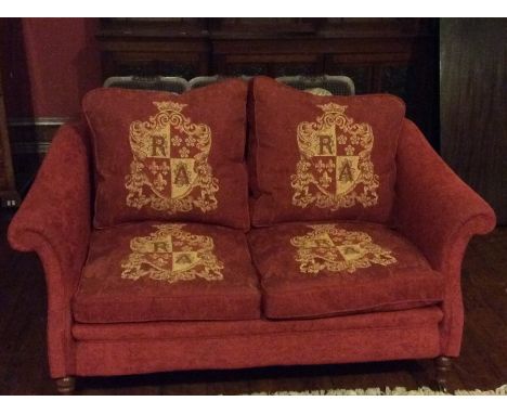 An early 20th Century red upholstered two seater sofa with heraldry cushions, scrolled arm rests, on turned supports on brass