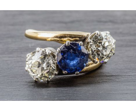 An 18ct gold, sapphire and diamond three stone cross over ring the central 6.6mm. round cut sapphire flanked by two round old