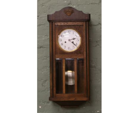 An Edwardian oak cased springer wall clock with white enamel dial, brass bezel, pendulum and key.