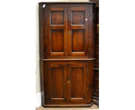 < A George III oak freestanding corner cupboard, the upper section with four fielded panel cupboard doors enclosing shelves t