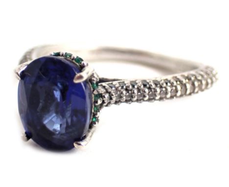 A dress ring, set with oval cut sapphire, surrounded by tiny emeralds to a raised shank setting with white metal set shoulder