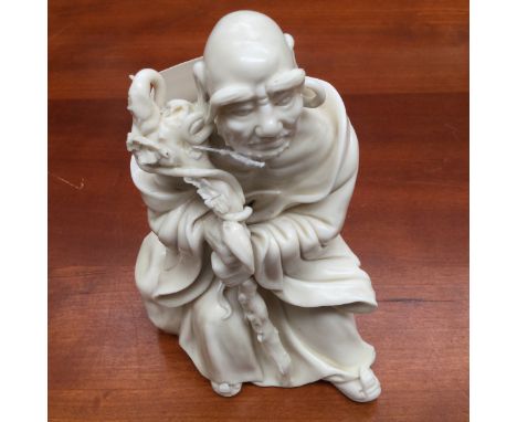 A Chinese blanc de chine of a seated Luohan, holding on to a gnarled root wood stick, bearing an artist square seal mark, bel