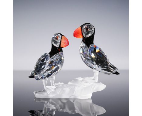 A charming Swarovski crystal figurine featuring a pair of puffins perched on a frosted crystal rock base. Designed by Michael