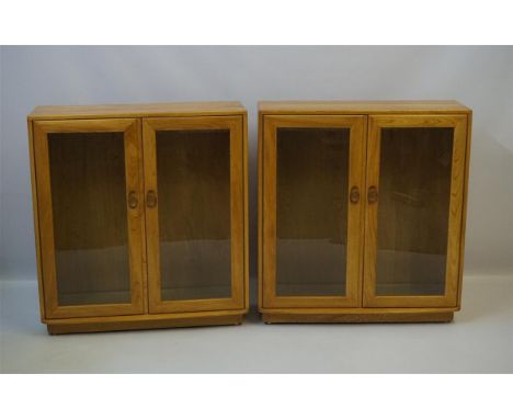 A pair of Ercol light elm bookcases, each having twin bevelled glazed doors enclosing two fixed shelves, raised on plinth bas