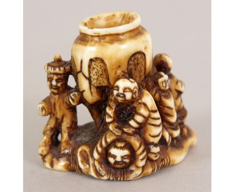 A SIGNED JAPANESE STAINED IVORY NETSUKE OF A GROUP OF BOYS, surrounding an urn supported on a rocky plinth, the base with an 