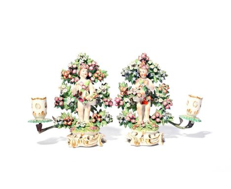 A pair of Bow candlestick figures of putti, c.1765-75, each modelled with a putto standing before flowering bocage and wearin