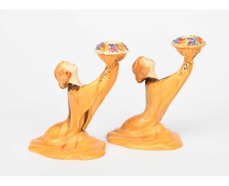 A pair of Clarice Cliff Bizarre Girl candlesticks, each modelled as a woman offering up a floral sconce, painted in colours p