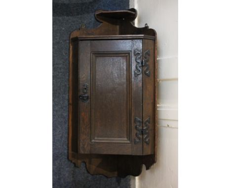 An oak hanging corner wall cupboard with small shelf surmount above panel door enclosing shelf, with decorative cast iron hin