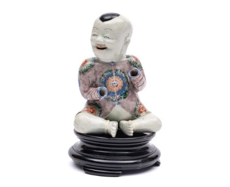 A Chinese famille verte seated figure of a smiling boy: wearing an aubergine coloured jacket decorated with floral roundels i