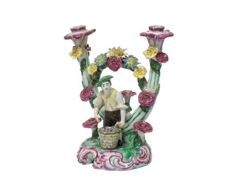 A French faience figural candlestick, 18th century, modelled as a young man standing with a basket of fruit at his feet betwe