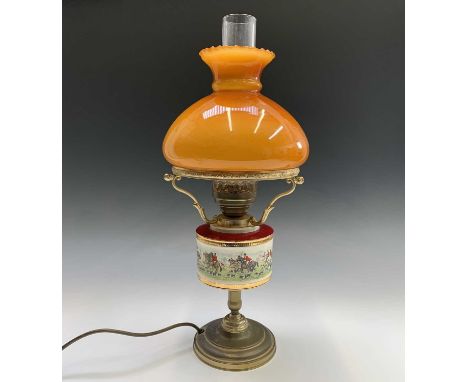 A brass effect oil lamp, the ceramic reservoir printed with a hunting scene, with amber glass shade. Overall height including