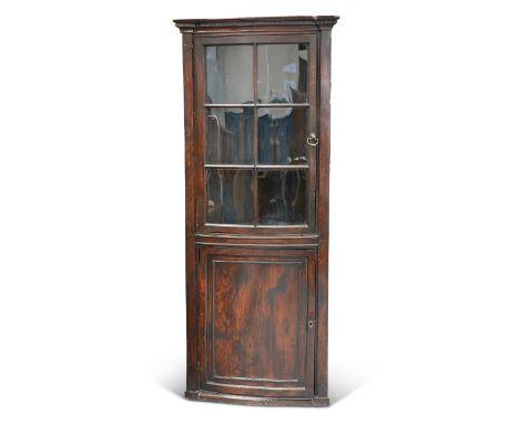 A GEORGE III ELM BOW-FRONT FLOOR-STANDING CORNER CUPBOARD, with moulded cornice above a glazed door with brass axe-head handl
