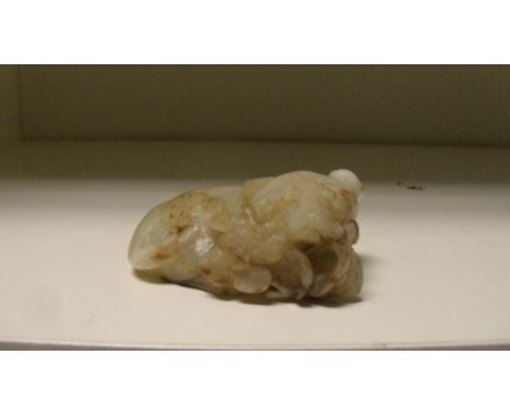 A jade carving of a cat, the grey green stone with brown inclusions depicting the feline with a flowering leaf spray in its m