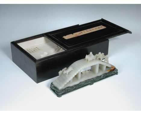 An ebony boxed nephrite jade bridge scene on spinach green stand, the pale grey green stone carved with a horseman and two ca