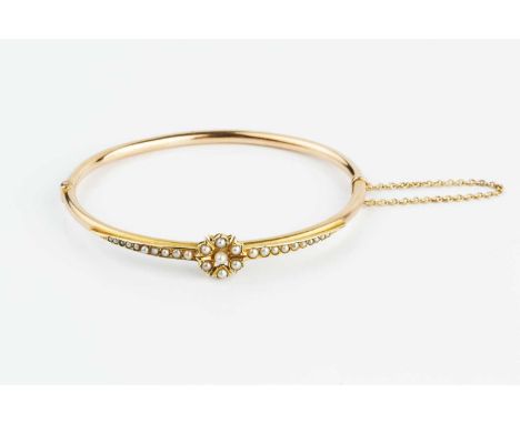 A late Victorian/Edwardian half pearl set bangle, of hinged oval form, applied with a half pearl set cluster between two simi