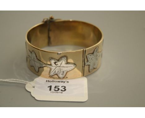 A Victorian mixed metal bangle, the wide band overlaid with a trailing frieze of ivy leaves, to a ruby set push button clasp