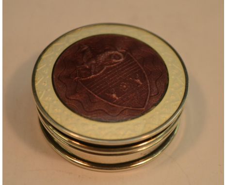 A George V silver and enamel circular novelty pill box, push-fitting cover decorated with an armorial, in tones of mauve and 