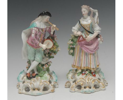 A Derby pair of Patch Mark figure, of a Musicians, she stands playing the triangle, wearing feathers in her hair, striped ski