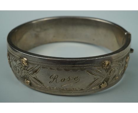A late 19th / early 20th Century white metal hinged bangle, engraved, faced with yellow metal foliate ornament and bearing th