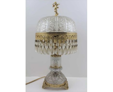 AN ORNATE GLASS &amp; BRASS TABLE LAMP, the shade with cherub finial and tear droppers, on shell foot base, 43cm high 