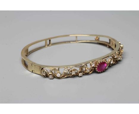 A STIFF HINGED BANGLE, the open upper section centred by an oval facet cut ruby, in scrolling foliage set with sixteen small 