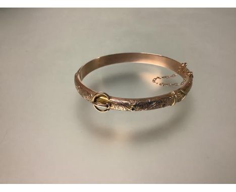 A 9ct gold hinged bangle, early 20th century, of belt and buckle form, foliate engraved, with safety chain. Internal diameter