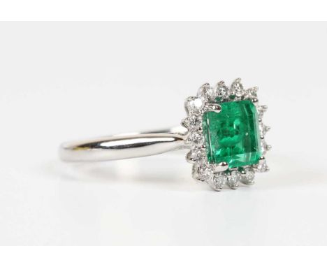 A platinum, emerald and diamond cluster ring, claw set with the cut cornered rectangular step cut emerald within a surround o