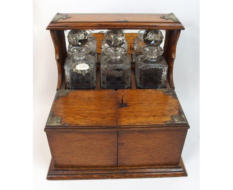 An Edwardian oak three tantalus/smokers cabinet of unusual form the three bottle decanter raised above a long drawer, the fro