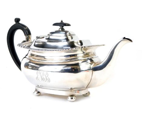 A George V silver teapot, with shell capped reeded borders, with an ebonised knop and handle, on bun feet bearing the crest E