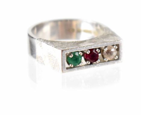 A 1970s modern 18ct white gold ring, channel set with a diamond, ruby and emerald, to a textured head and polished shank, Lon