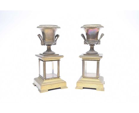 A pair of brass campana urns, on integral pedestals, with bevelled glass panels and shaped plinths, some damage/wear, 25cm hi