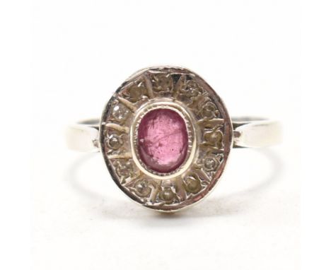 A hallmarked 9ct white gold, ruby and diamond halo ring. The ring having a central bezel set oval cut ruby encompassed by a c