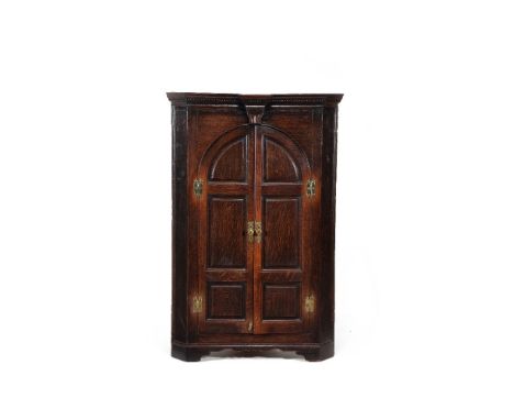 A George III oak standing corner cupboard, circa 1770, with dentil cornice and waved frieze above a pair of arched panel door