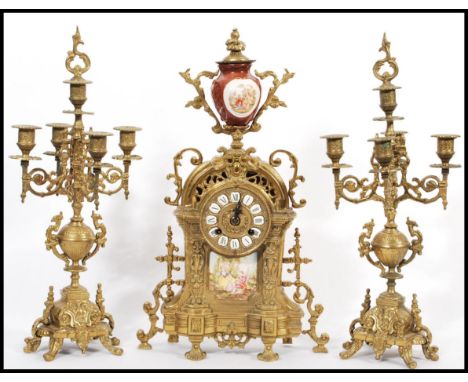 A vintage 20th century German gilt brass mantel clock and garniture set. The clock with red ceramic urn and painted scenes on