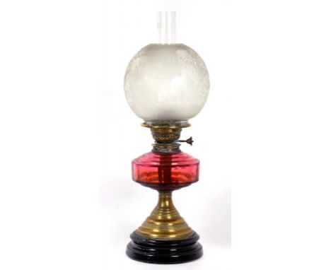 A LATE VICTORIAN BRASS OIL LAMP WITH FACETED CRANBERRY GLASS FOUNT AND BRASS DUPLEX BURNER, ON BLACK GLAZED EARTHENWARE FOOT,