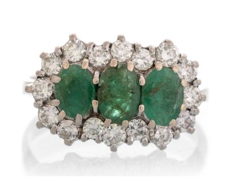 EMERALD AND DIAMOND CLUSTER RING, IN UNMARKED WHITE GOLD OR PLATINUM  with three oval emeralds in a surround of sixteen round